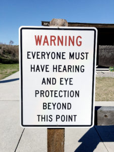 Vertical white metal sign with a thin black border attached to a wooden post at an outdoor shooting range. The sign has WARNING in big red letters followed by EVERYONE MUST HAVE HEARING AND EYE PROTECTION BEYOND THIS POINT in capital black letters.