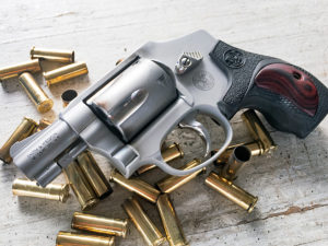 A silver Smith & Wesson .38 Special +P subcompact self-defense revolver with fancy Performance Center grips, lying on a wooden backdrop among a pile of spent .38 Special brass casings.